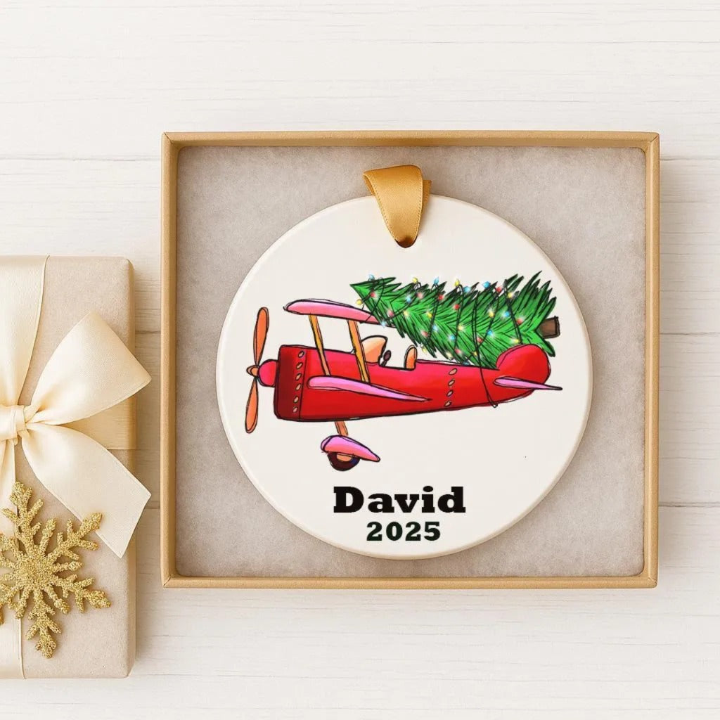 A white ornament with a red propeller airplane carrying a Christmas tree is displayed in a gold frame.