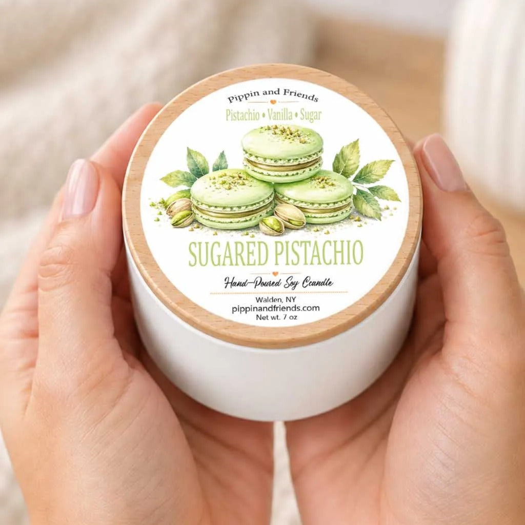 A person's hands holding a white container with a label that features illustrations of green pistachio macarons and leaves.
