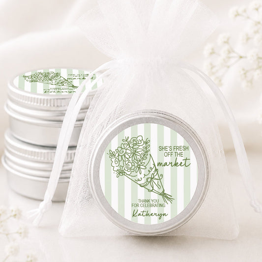A small round silver tin wrapped in a sheer white organza drawstring bag, with a printed label that reads 'She's Fresh Off the Market' and shows a green botanical illustration. More matching silver tins are stacked in the background on a soft white surface.