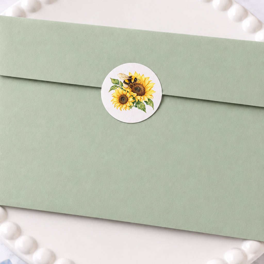 Close-up of a sage green envelope sealed with a round sticker featuring watercolor sunflowers and a bumblebee, styled on a white decorative plate with soft blue floral accents.