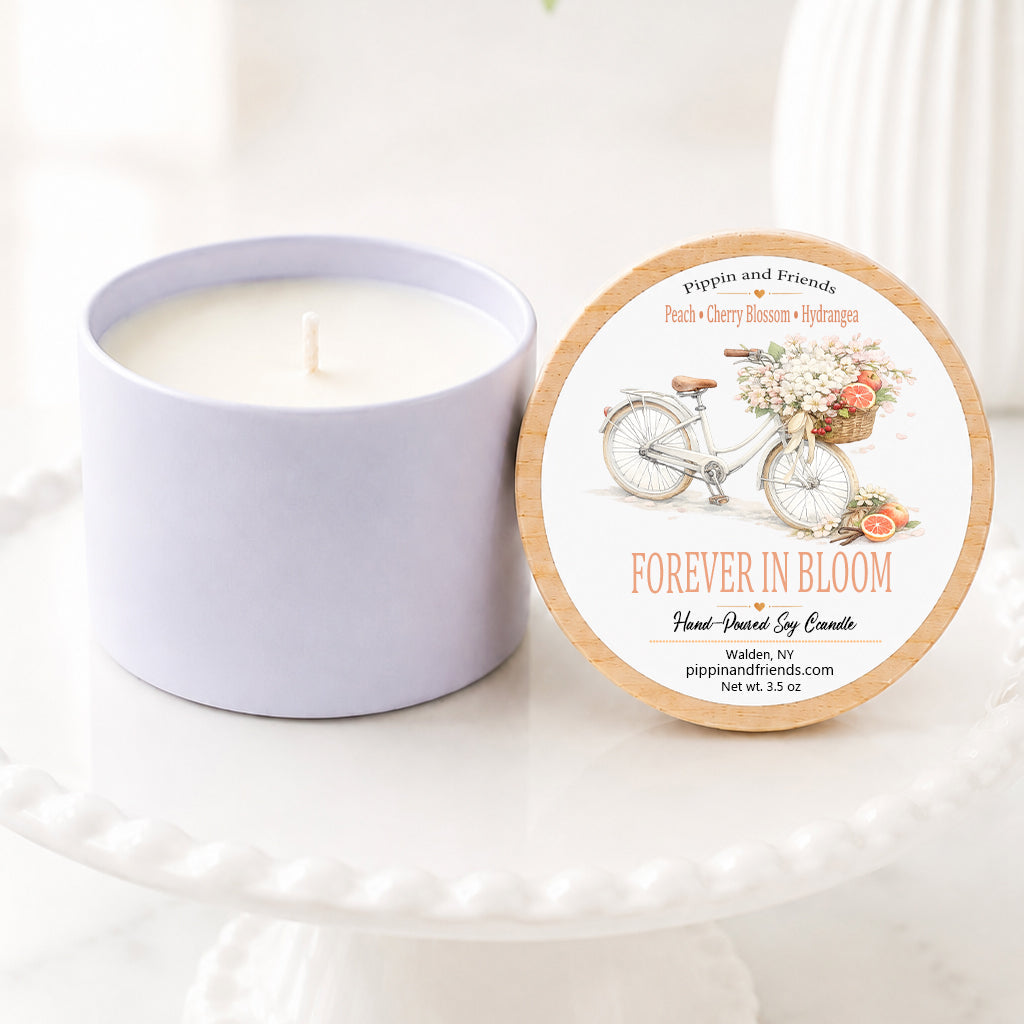 A soy candle in a matte cup container sits next to its round lid on a white pedestal. The lid features a vintage bicycle illustration and the scent name 'FOREVER IN BLOOM'.