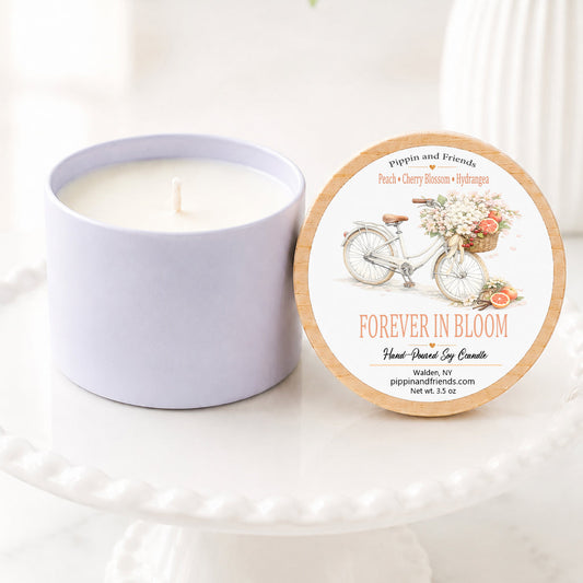 A soy candle in a matte cup container sits next to its round lid on a white pedestal. The lid features a vintage bicycle illustration and the scent name 'FOREVER IN BLOOM'.