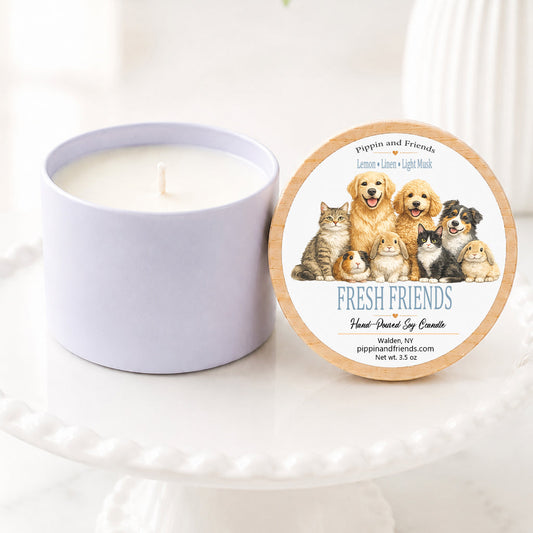A  soy candle in a white jar sits on a white pedestal next to its round lid, which features an illustration of various pets and the text 'Fresh Friends. The background is a bright, softly blurred indoor setting with a white vase of flowers.