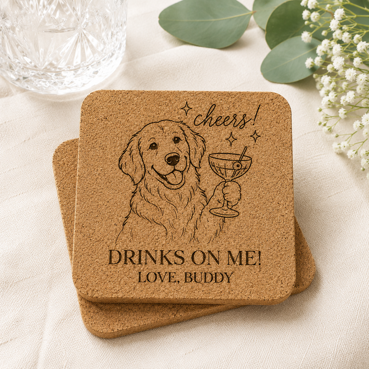 Personalized cork coaster featuring a laser-engraved golden retriever holding a cocktail glass with the phrase "Cheers! Drinks On Me! Love, Buddy," displayed in a light and airy setting with eucalyptus greenery, baby's breath flowers, and a crystal glass.