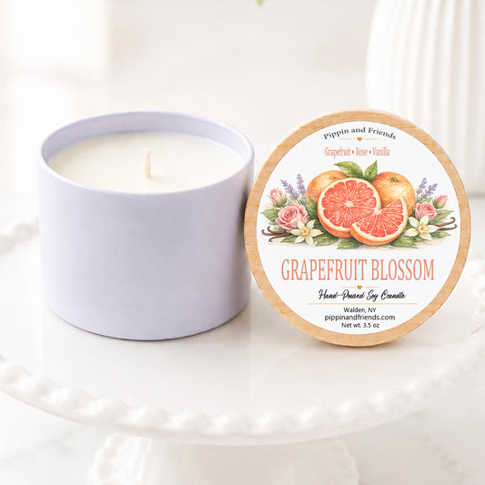 Grapefruit scented hand-poured soy candle in a matte white vessel with natural wooden lid displayed upright beside it, featuring a watercolor grapefruits and floral label. The candle sits on a white scalloped pedestal against a bright, airy background with soft white flowers in a vase, creating a clean, cozy woodland-inspired aesthetic.