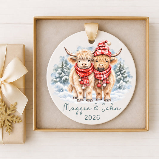 Personalized Highland cow couple Christmas ornament featuring two fluffy watercolor Highland cows wearing red plaid scarves, one with a matching winter hat, standing in a snowy evergreen forest. Custom names Maggie and John with year 2026, printed on a round ceramic ornament with satin ribbon, displayed in a keepsake gift box.