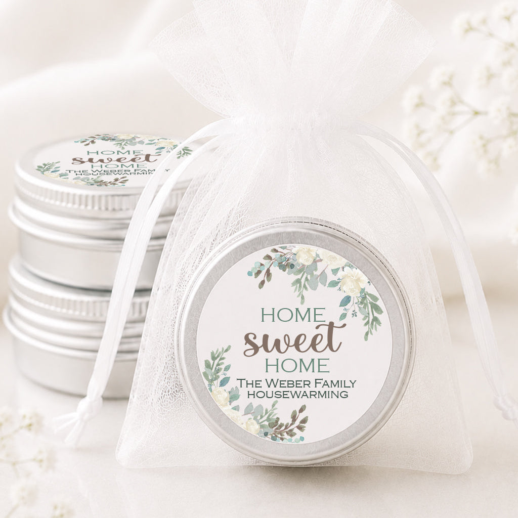 A round silver tin wrapped in a sheer white organza bag with a ribbon, featuring a floral label that reads 'HOME sweet HOME THE WEBBER FAMILY HOUSEWARMING.' Another similar tin sits behind it on a soft white fabric background with delicate white flowers around it.