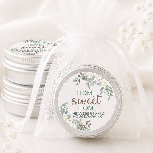 A round silver tin wrapped in a sheer white organza bag with a ribbon, featuring a floral label that reads 'HOME sweet HOME THE WEBBER FAMILY HOUSEWARMING.' Another similar tin sits behind it on a soft white fabric background with delicate white flowers around it.