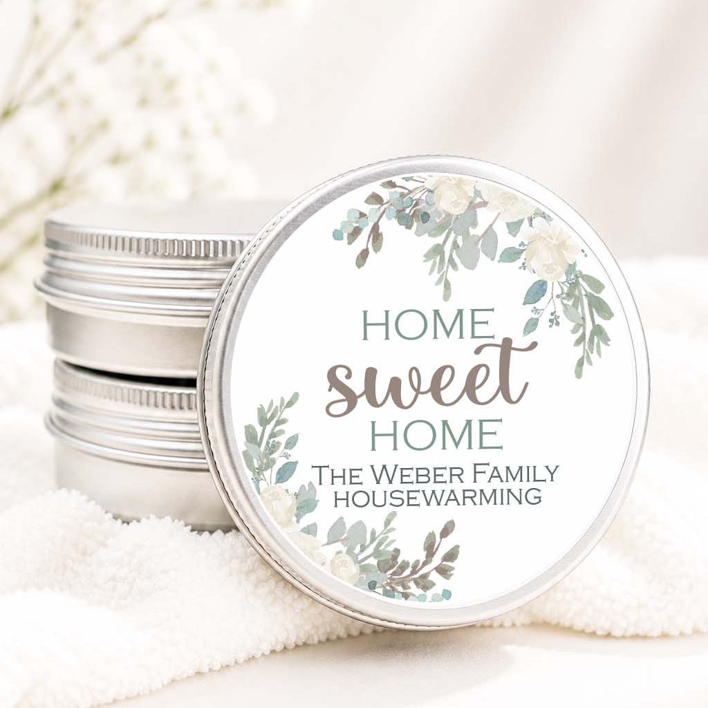 A stack of round silver metal tins with one tin open and its lid leaning against it. The lid has a white printed label with a floral wreath design and the text 'HOME sweet HOME THE WEBER FAMILY HOUSEWARMING.