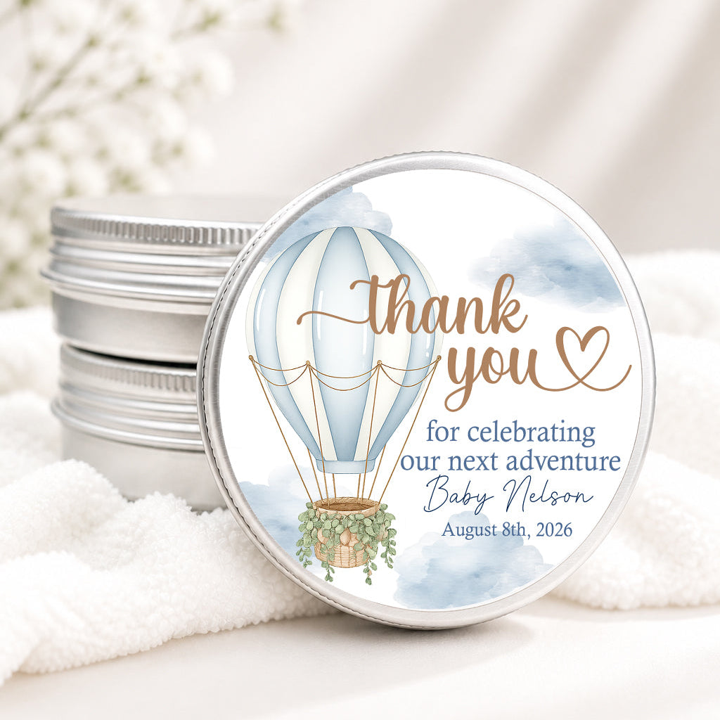 Round silver metal gift tin with a printed hot air balloon thank you label, shown in front of a stack of matching tins on soft white fabric with blurred white flowers in the background.