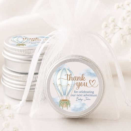A round silver tin wrapped in a sheer white organza drawstring bag with a printed circular label that reads 'thank you for celebrating our next adventure Baby John' with a hot air balloon illustration. More matching silver tins are stacked in the background on a soft white fabric surface.