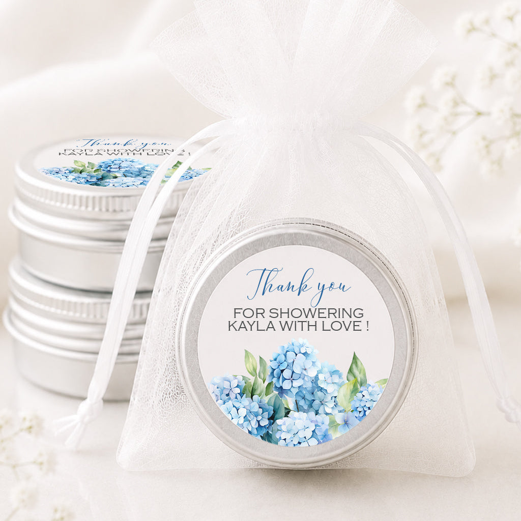 A round silver tin with a personalized blue hydrangea thank you label, tied in a sheer white organza pouch, with other matching silver tins stacked in the background.