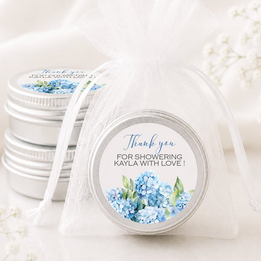 A round silver tin with a personalized blue hydrangea thank you label, tied in a sheer white organza pouch, with other matching silver tins stacked in the background.
