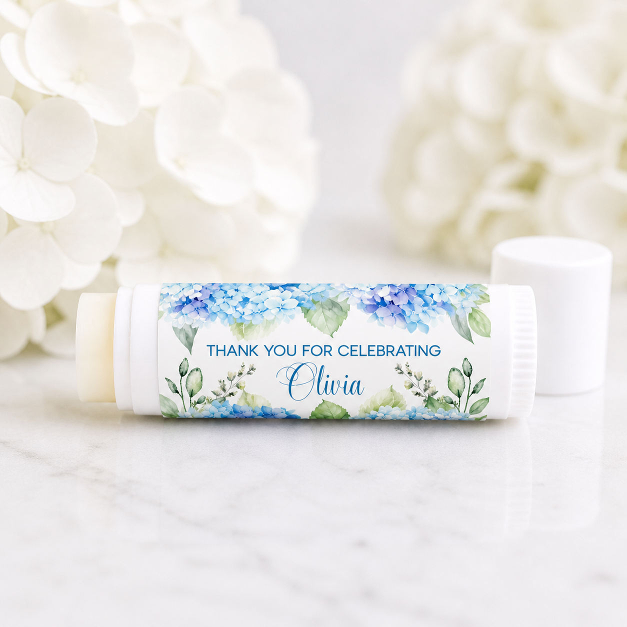 Hydrangea lip balm favor with personalized waterproof label reading “Thank You for Celebrating Olivia,” displayed on a white tube with soft white hydrangea flowers in the background.