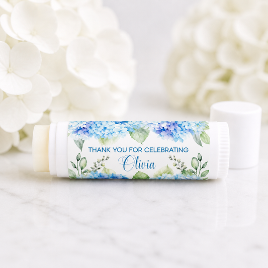 Hydrangea lip balm favor with personalized waterproof label reading “Thank You for Celebrating Olivia,” displayed on a white tube with soft white hydrangea flowers in the background.