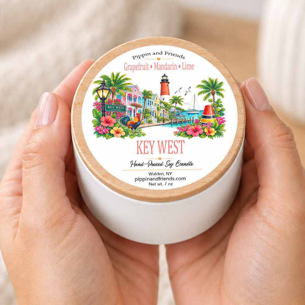 A person holds a white jar candle with a light wood lid in both hands. The lid features a colorful tropical illustration and the text 'KEY WEST' and 'Pipern and Friends'.
