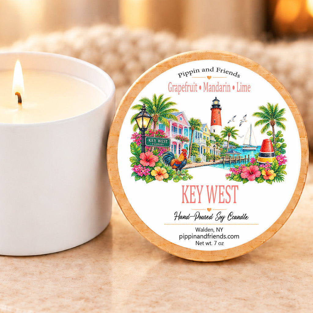 A round scented soy candle tin labeled 'Pipelin and Friends Key West Hand-Painted Soy Candle' sits on a stone surface beside a lit white candle jar. The lid features a colorful illustration of a coastal building, lighthouse, palm trees, and flowers.
