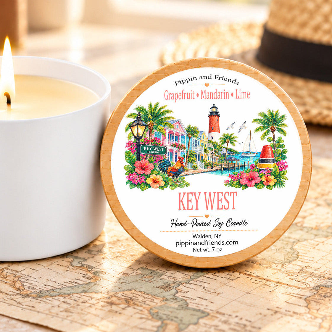 A round scented soy candle tin labeled 'KEY WEST' by Pippin and Friends sits on a wooden surface beside a lit white candle. The lid features a colorful tropical harbor illustration with palm trees, flowers, and a lighthouse.
