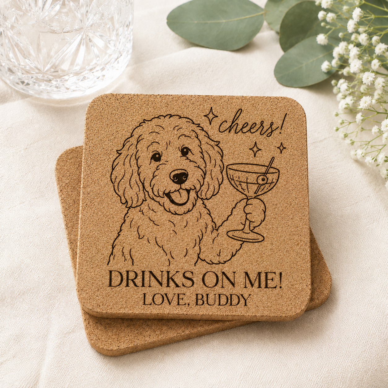 Personalized cork coaster featuring a laser-engraved Labradoodle holding a cocktail glass with the phrase "Cheers!" and custom text reading "Drinks On Me! Love, Buddy," displayed on a soft neutral fabric with eucalyptus greenery, baby's breath flowers, and a crystal glass in a light and airy setting.