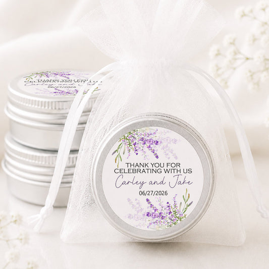 A small round silver favor tin wrapped in a sheer white organza bag, with a printed round label that reads 'THANK YOU FOR CELEBRATING WITH US Carley and Jake 06/27/2026' and features purple floral artwork. More matching favor tins are stacked in the background on a soft white surface.