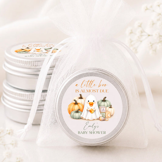 A small round silver tin wrapped in a sheer white organza bag, with a printed circular label reading 'a little bee is almost due' and 'Emily's baby shower' with autumn-themed illustrations. More matching silver tins are stacked in the background on a soft white surface.