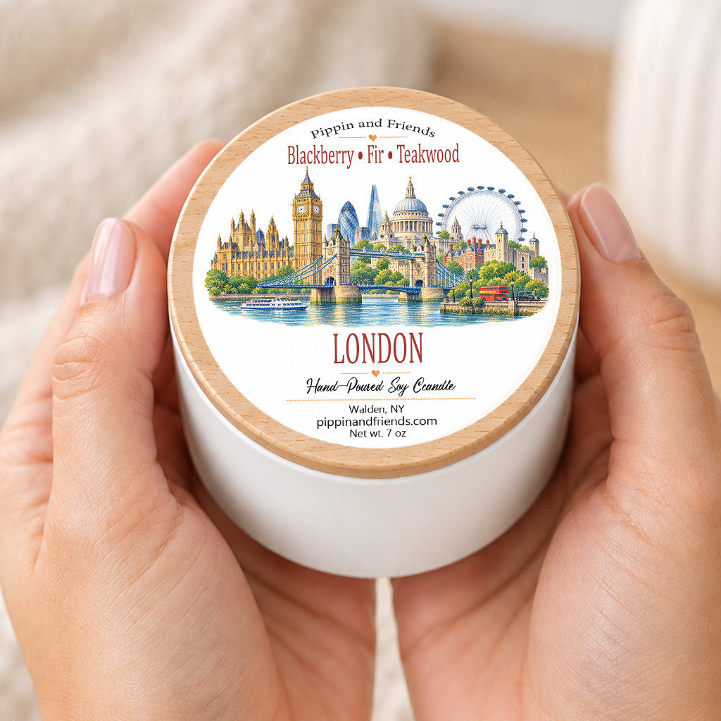 A person's hands holding a white jar candle with a light wood lid. The lid features a colorful illustration of London landmarks, including Big Ben, St.