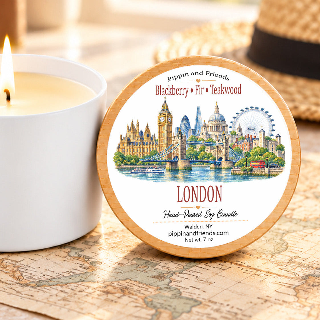 A round wooden-lidded candle tin labeled 'Pippin and Friends London Blackberry Fir • Teakwood' sits on a vintage-style world map next to a lit white candle. A straw hat is blurred in the background.