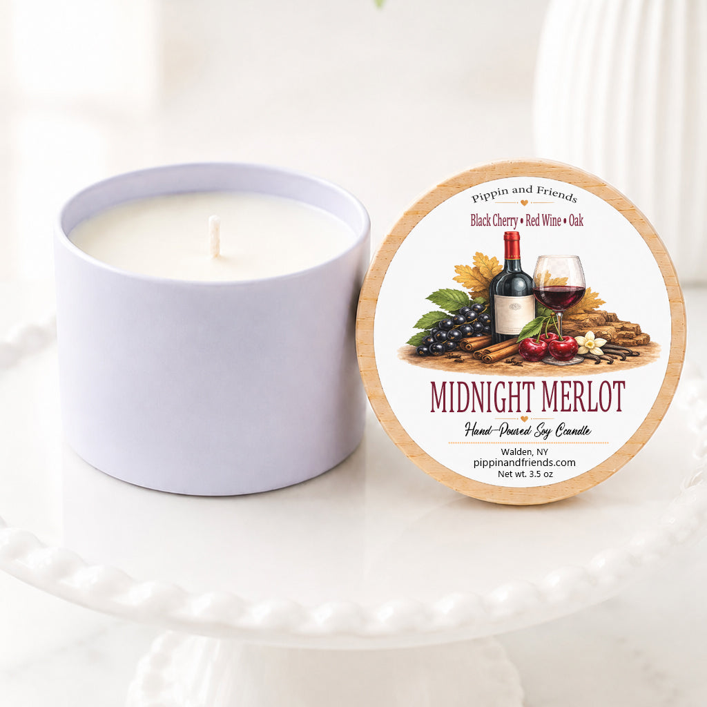 A  soy candle in a matte container sits next to its round lid on a white pedestal. The lid features a wine-themed label with the scent name 'Midnight Merlot' and the brand 'Pippin and Friends'.