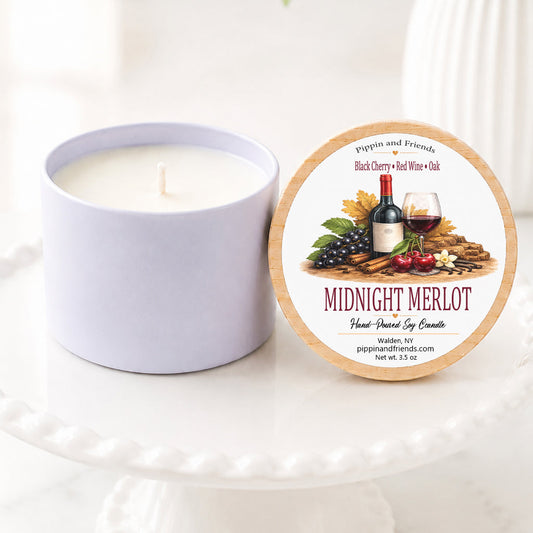 A  soy candle in a matte container sits next to its round lid on a white pedestal. The lid features a wine-themed label with the scent name 'Midnight Merlot' and the brand 'Pippin and Friends'.