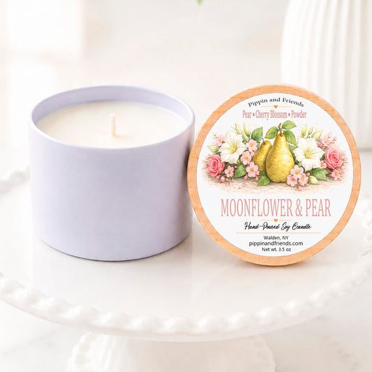 A soy candle jar sits beside its round lid on a white pedestal. The lid features a floral illustration with a pear and the scent name 'Moonflower & Pear.