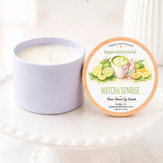 A soy candle in a matte jar sits next to its circular lid on a white pedestal. The lid features a wood-grain border and a colorful illustration of matcha green tea and lemon slices, with the scent name 'MATCHA SUNRISE' and brand 'pippin and friends' visible.