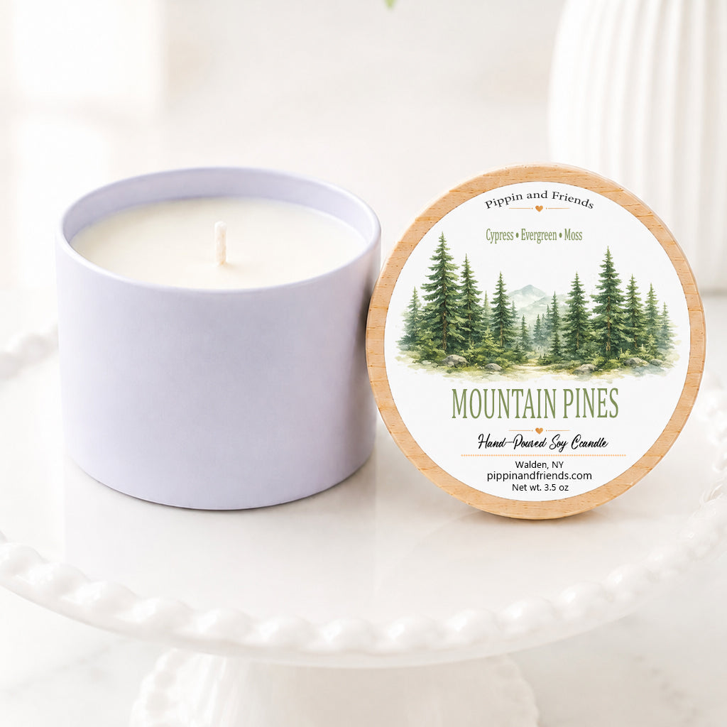 Mountain Pines hand-poured soy candle in a matte white vessel with natural wooden lid displayed upright beside it, featuring a watercolor evergreen forest label. The candle sits on a white scalloped pedestal against a bright, airy background with soft white flowers in a vase, creating a clean, cozy woodland-inspired aesthetic.
