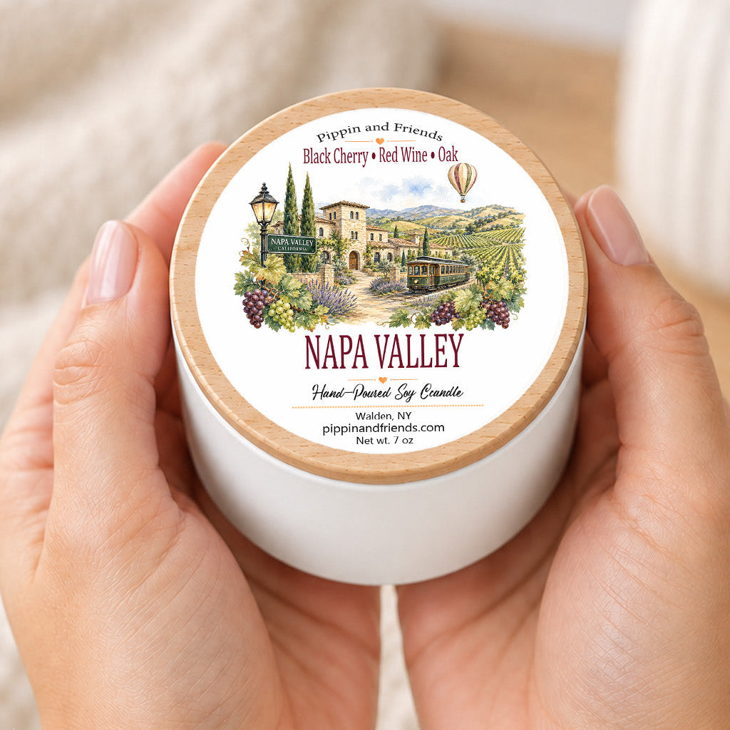 A person's hands holding a white jar candle with a light wood lid. The lid features a detailed illustration of a vineyard labeled 'NAPA VALLEY' with the scent 'Pippin & Friends'.