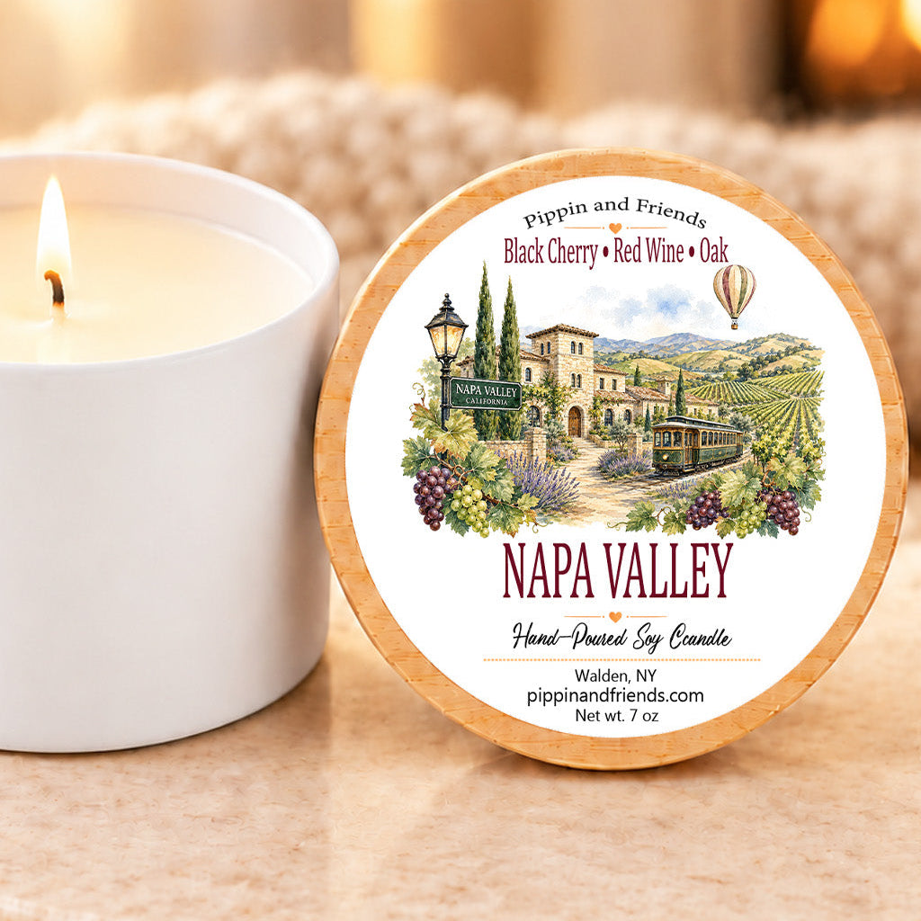 A round wooden scented candle lid labeled 'NAPA VALLEY' with an illustrated vineyard scene, placed beside a lit white candle on a stone surface in a warm indoor setting.