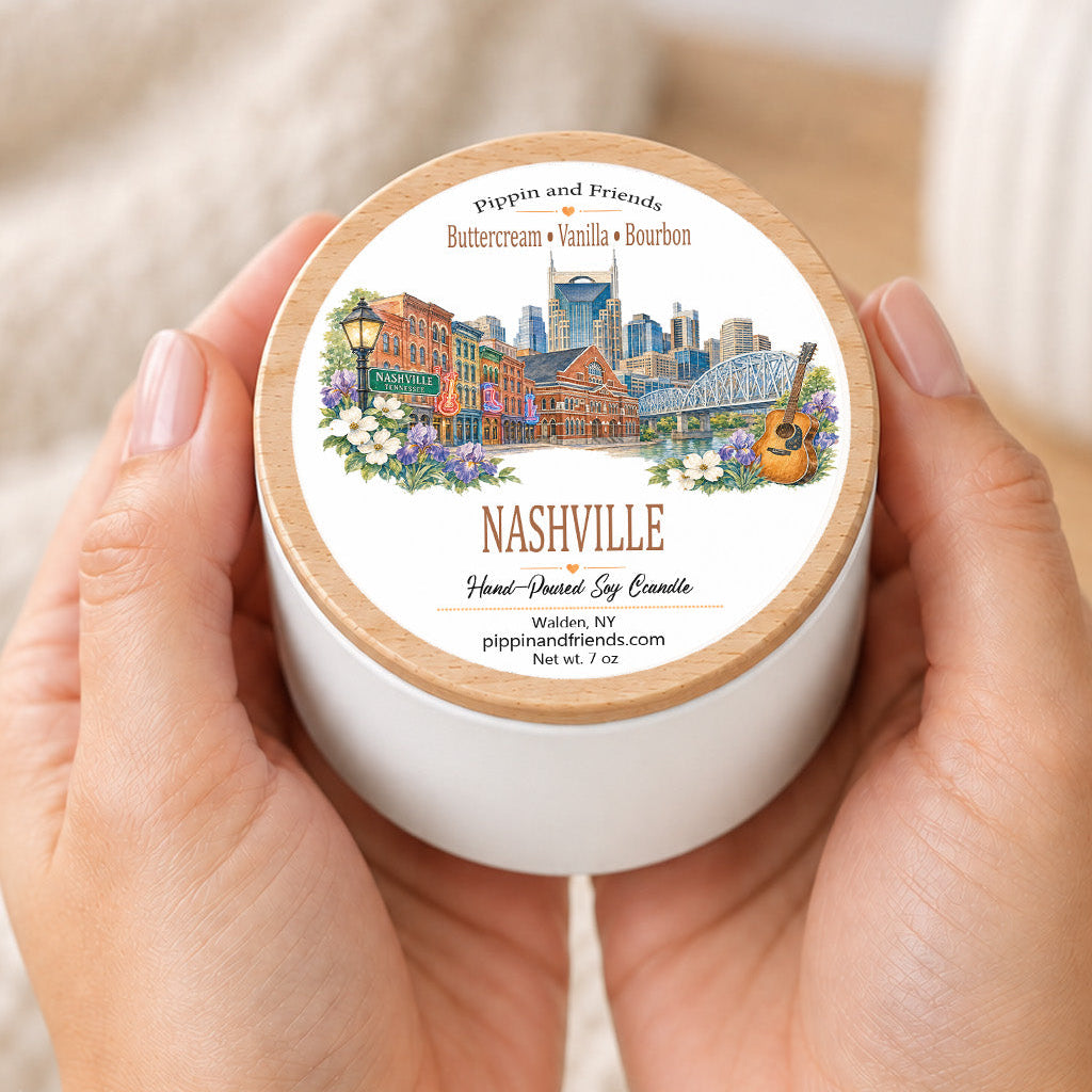 A person's hands holding a white jar candle with a light wood lid. The lid features a colorful illustration of a Nashville cityscape with the text 'Pipkin and Friends' and 'Nashville Hand-Soy Candle'.