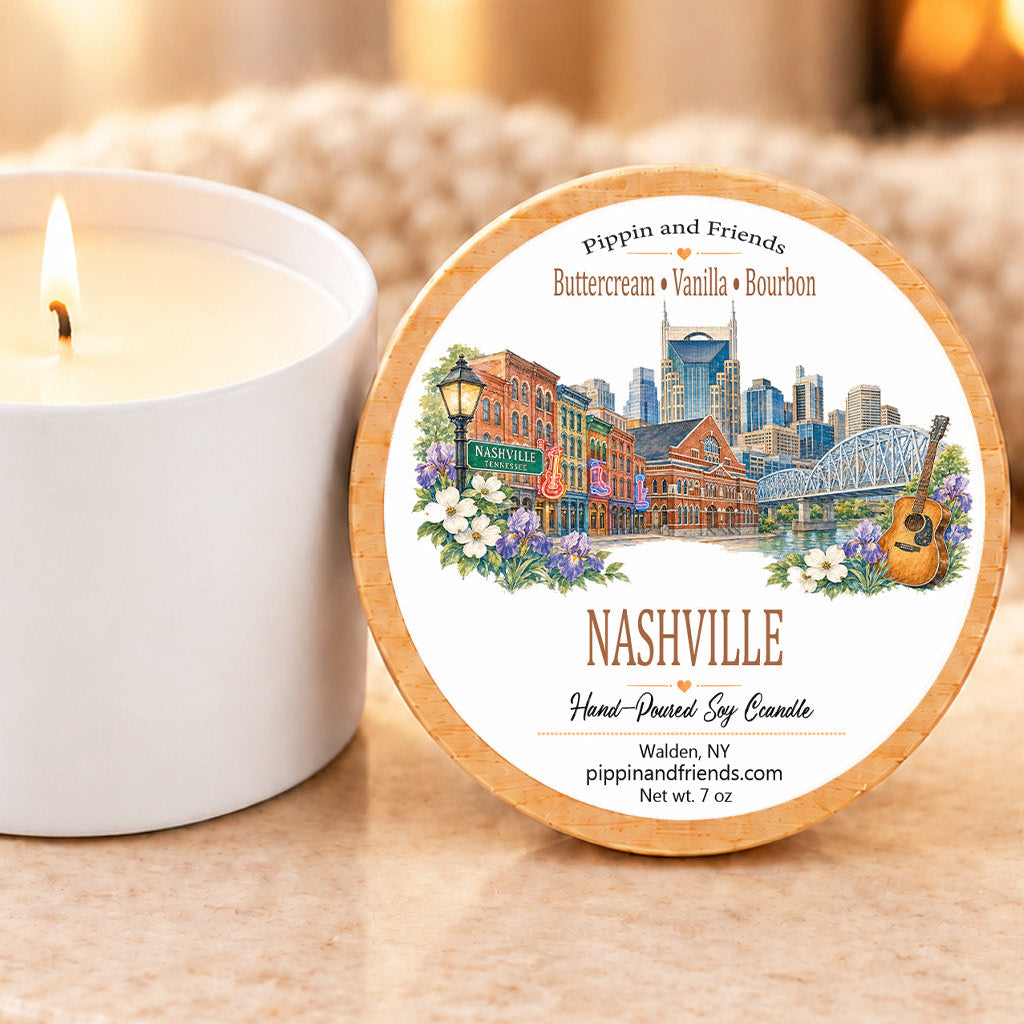 A round wooden lid with a colorful Nashville cityscape illustration and the text 'Pippin and Friends Buttercream + Vanilla + Bourbon' sits beside a lit white candle on a stone surface in a warm indoor setting.