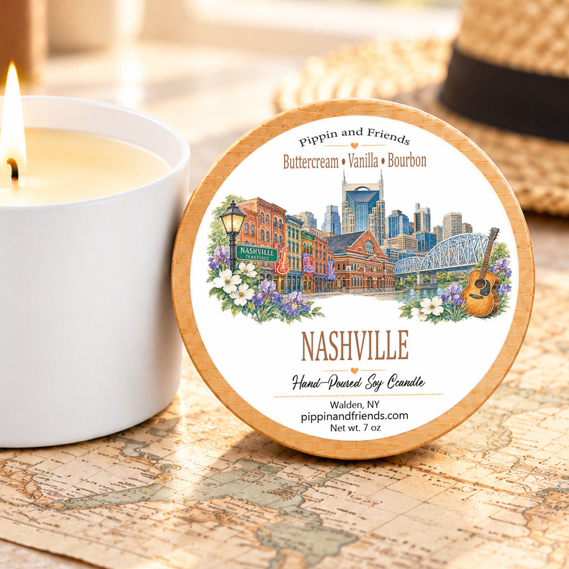 A round scented soy candle tin labeled 'Nashville' with a colorful city skyline illustration sits on a vintage map next to a lit white candle. A straw hat is blurred in the background.
