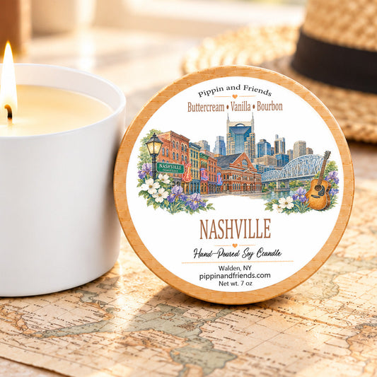 A round scented soy candle tin labeled 'Nashville' with a colorful city skyline illustration sits on a vintage map next to a lit white candle. A straw hat is blurred in the background.