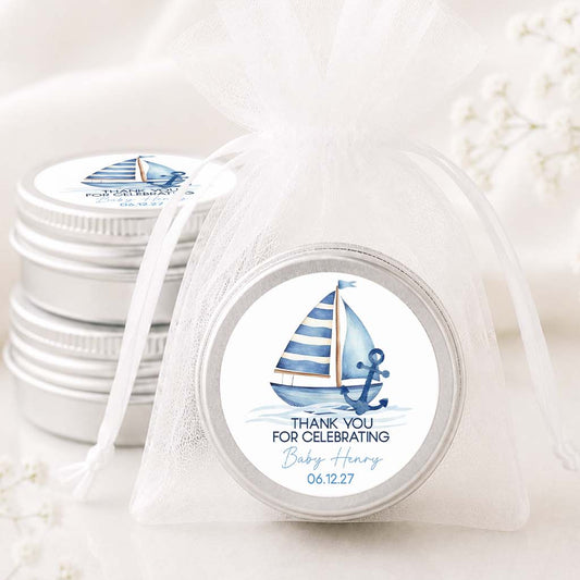 A round silver tin of baby keepsakes wrapped in a sheer white organza drawstring bag, featuring a blue and white sailboat label that reads 'THANK YOU FOR CELEBRATING Baby Henry 06.12.