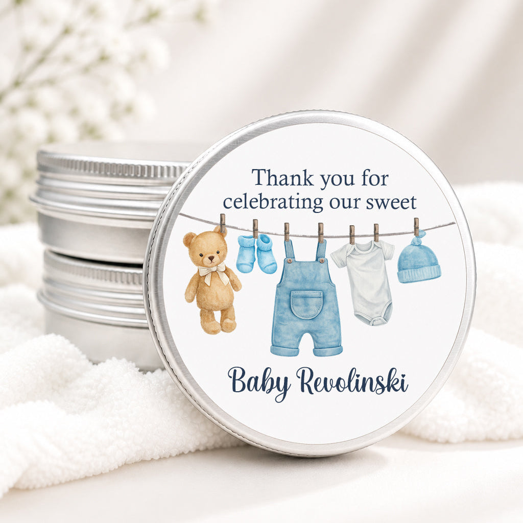 A round silver tin lid with a personalized baby shower favor design, featuring a watercolor teddy bear, blue overalls, socks, and a white onesie on a clothesline, with the text 'Thank you for celebrating our sweet Baby Revelinski'. The lid is shown resting on a white towel next to a stack of similar silver tins.
