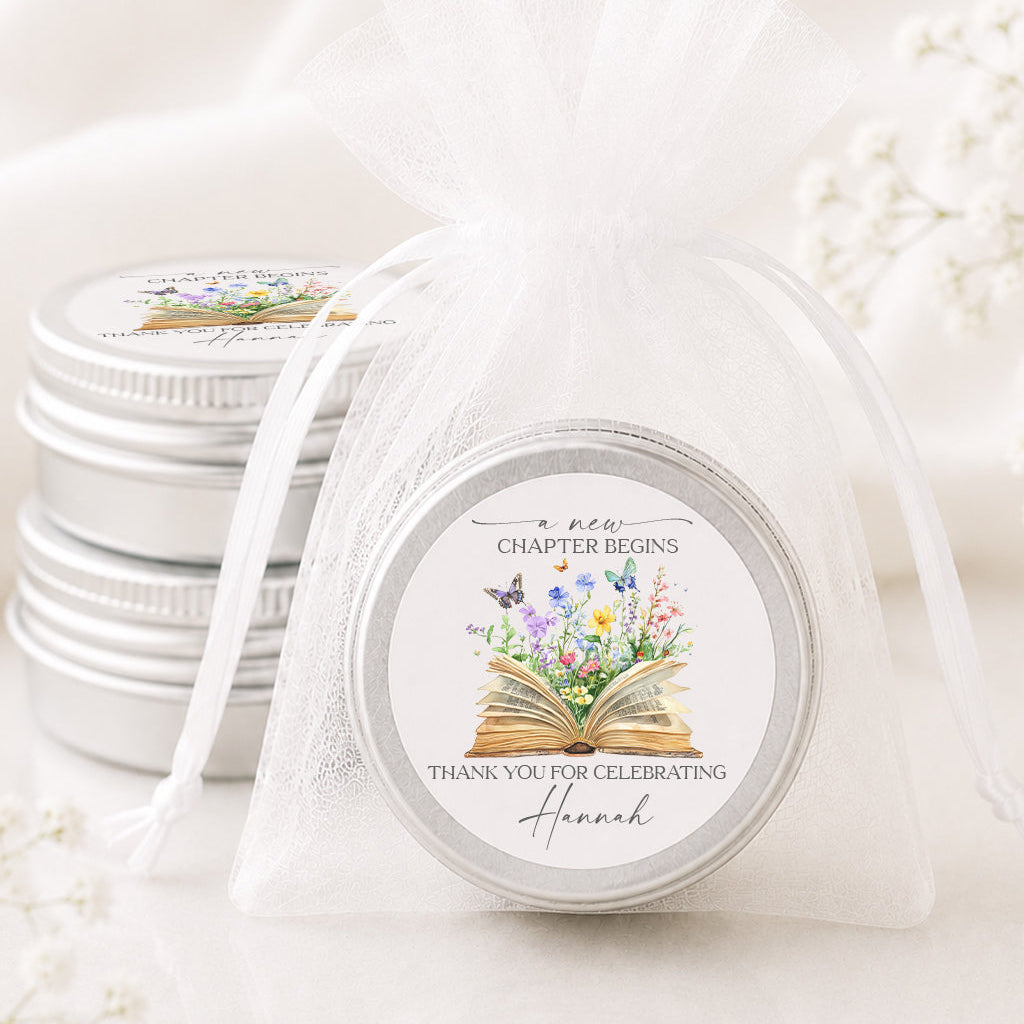 A round silver tin wrapped in a sheer white organza bag, with two more matching tins stacked in the background. The front tin lid features a floral and book illustration with the text 'A new chapter begins' and 'Thank you for celebrating Hannah'.
