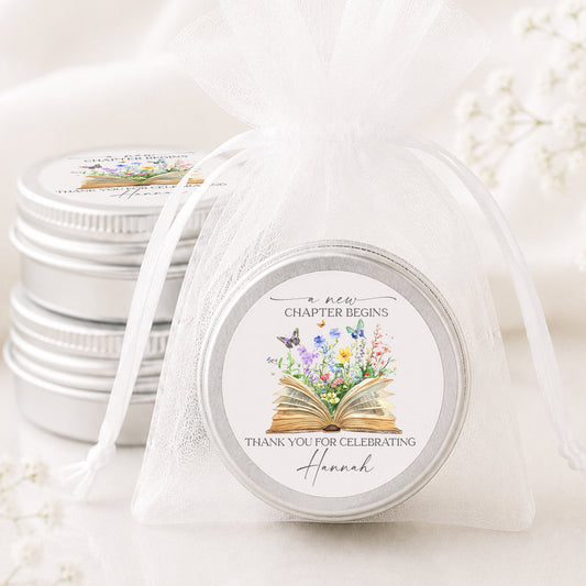 A round silver tin wrapped in a sheer white organza bag, with two more matching tins stacked in the background. The front tin lid features a floral and book illustration with the text 'A new chapter begins' and 'Thank you for celebrating Hannah'.