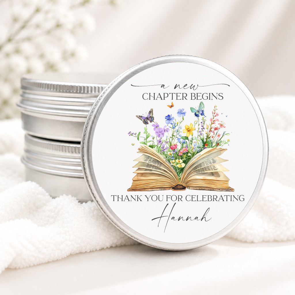 A round silver metal tin with a white lid printed with an illustration of an open book, flowers, and butterflies, resting on white fabric next to a stack of similar tins.