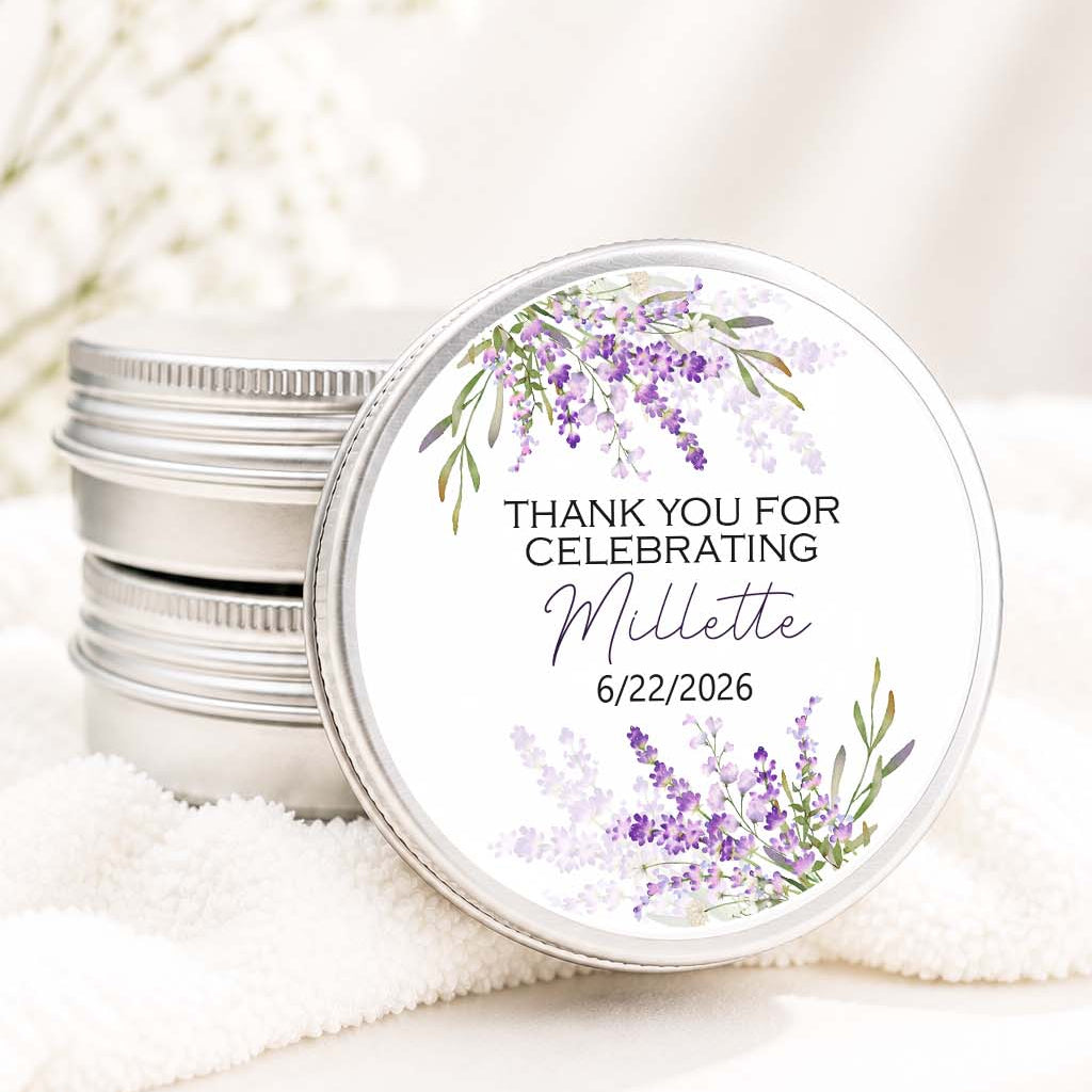 A stack of round silver metal tins with a single tin in front showing a white label printed with purple floral artwork and the text 'THANK YOU FOR CELEBRATING Millette 6/22/2026.' The tins are styled on a soft white towel with blurred white flowers in the background.