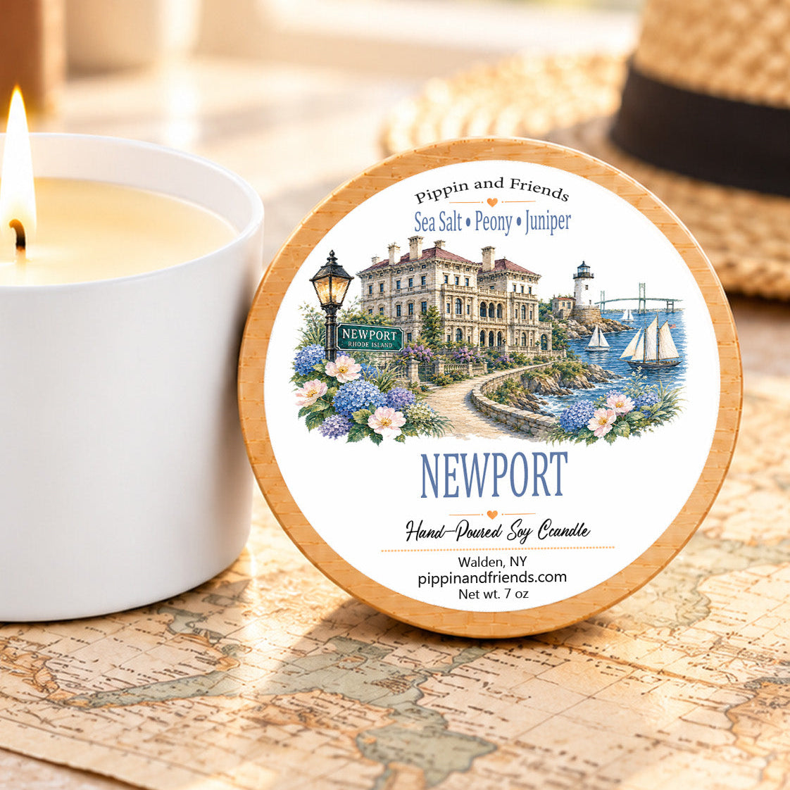 A round tin candle labeled Newport by Pippin and Friends sits on a vintage-style map next to a lit white candle jar. A straw hat is blurred in the background.