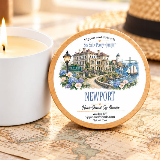 A round tin candle labeled Newport by Pippin and Friends sits on a vintage-style map next to a lit white candle jar. A straw hat is blurred in the background.