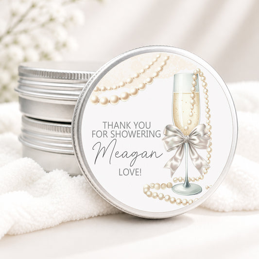 Personalized bridal shower candle favor in a silver tin featuring a champagne flute with a satin bow, elegant pearl accents, and custom event details, displayed on a soft white background.
