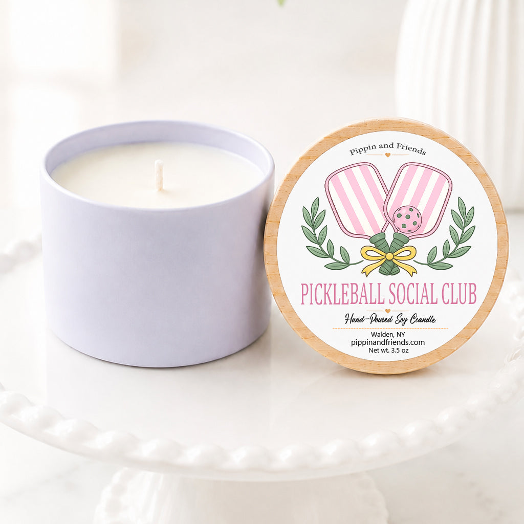 A soy candle in a matte cup sits next to its round lid on a white pedestal. The lid features a label for 'Pickleball Social Club' by 'Pippin and Friends', decorated with pink marshmallows, green leaves, and a gold bow.