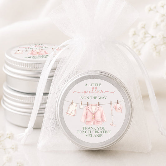 A round silver tin wrapped in a sheer white organza drawstring bag, with a printed circular label that reads 'A LITTLE butter IS ON THE WAY THANK YOU FOR CELEBRATING MELANIE' and shows pink baby clothes hanging on a line. More matching silver tins are stacked in the background on a soft white surface with draped fabric and small white flowers.
