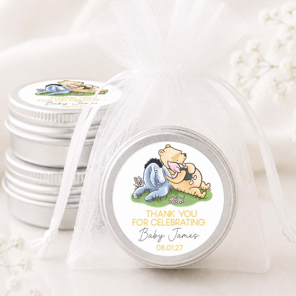 A round silver favor tin wrapped in a sheer white organza bag, with a custom round label featuring Winnie the Pooh and Eeyore. A stack of similar favor tins sits behind it on a soft white fabric background with delicate white floral accents.